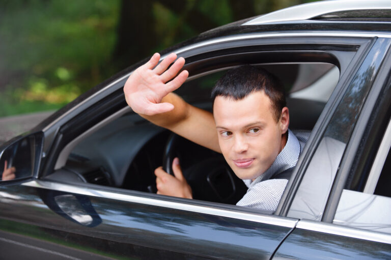 Portrait of young man driving car and greeting somebody with han