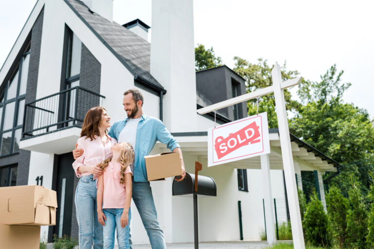 bearded man holding box and standing with wife and daughter near house and board with sold letters