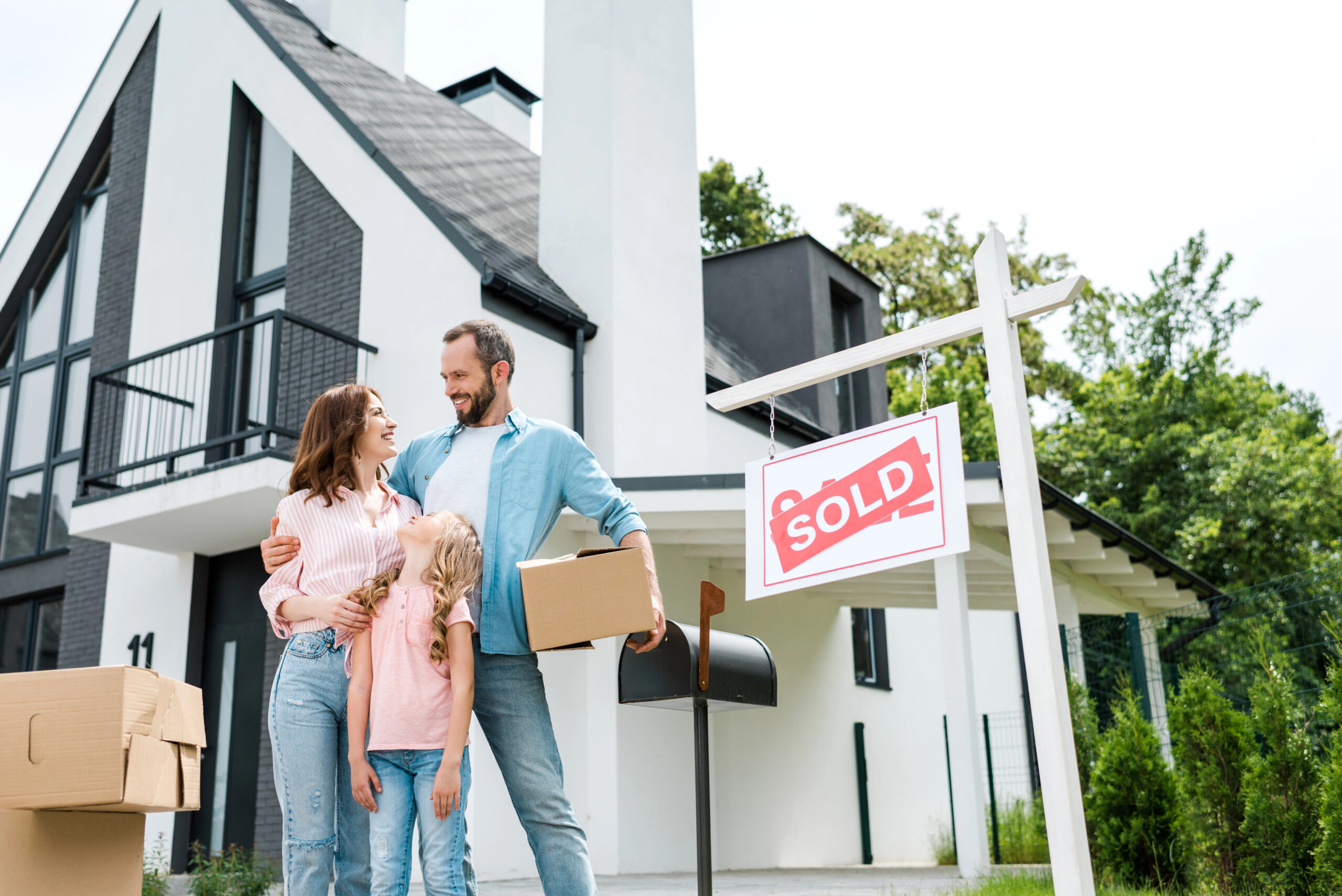 bearded man holding box and standing with wife and daughter near house and board with sold letters